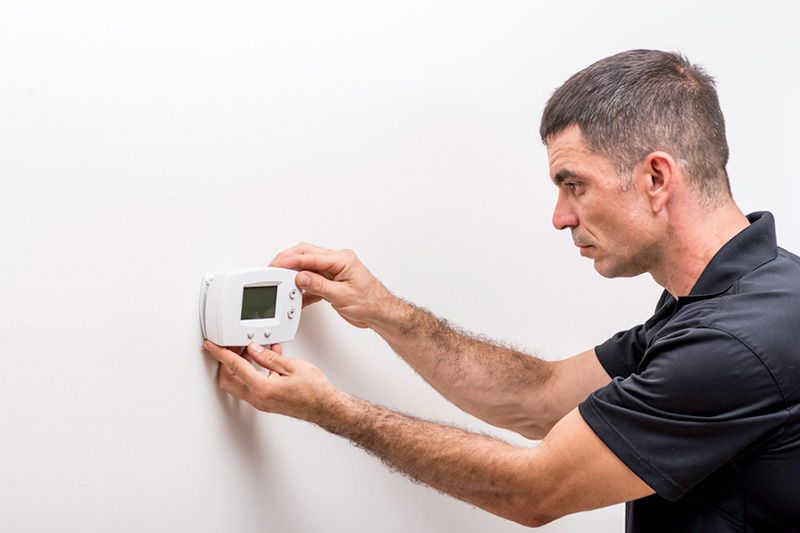 Tips to Troubleshoot Your Thermostat. Image shows man with short grey hair wearing a black t-short troubleshooting thermostat that is mounted on a white wall.